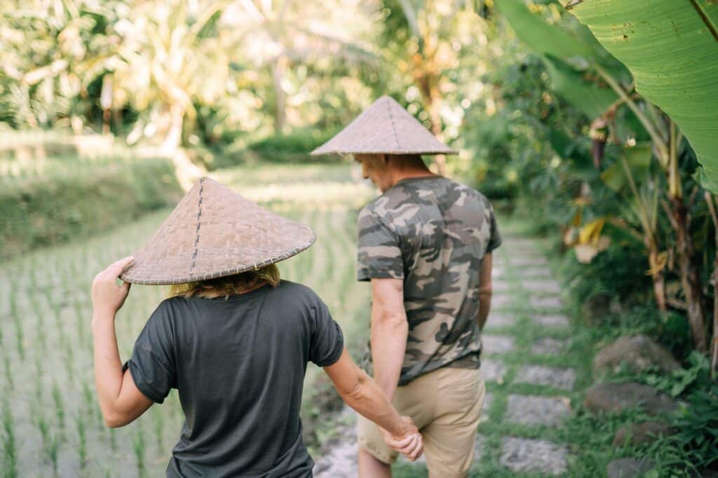 Un couple de voyageurs portant des chapeaux traditionnels coniques se tenant la main lors d'une promenade romantique dans les rizières à Bali avec Heaven Collection