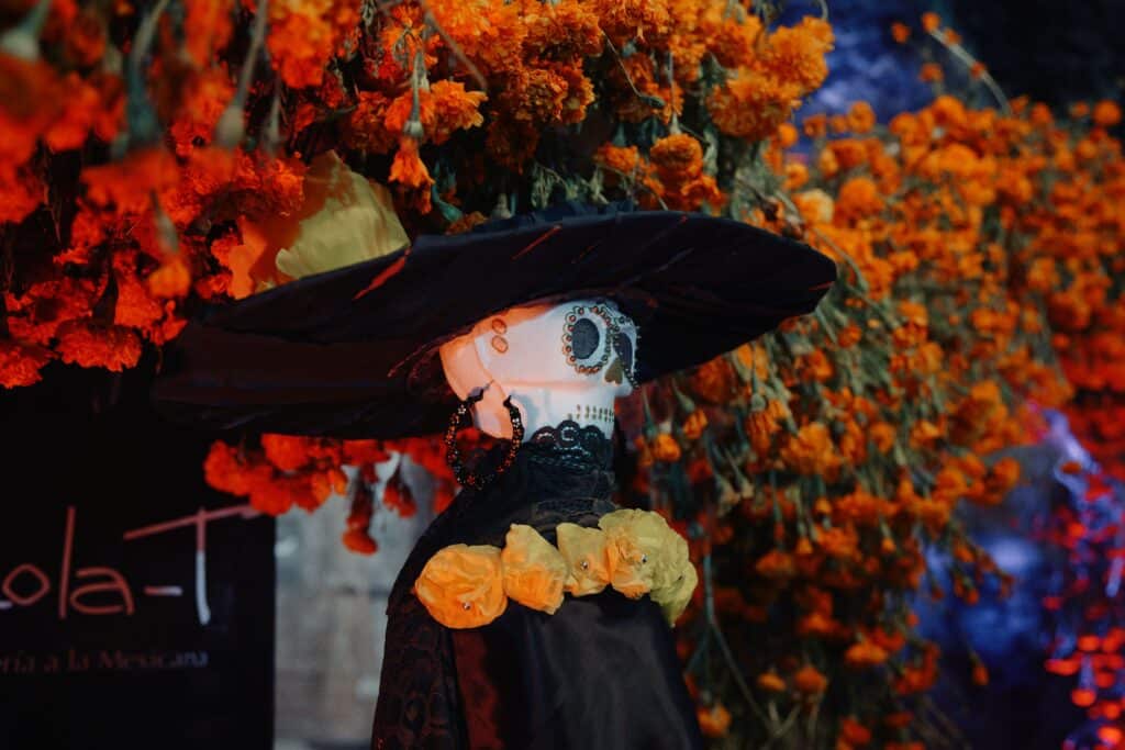 Buste de la Catrina, élégante figure squelettique vêtue de noir, coiffée d'un grand chapeau noir et de fleurs jaunes, se détachant sur un mur de fleurs d'œillets d'Inde orange vif.