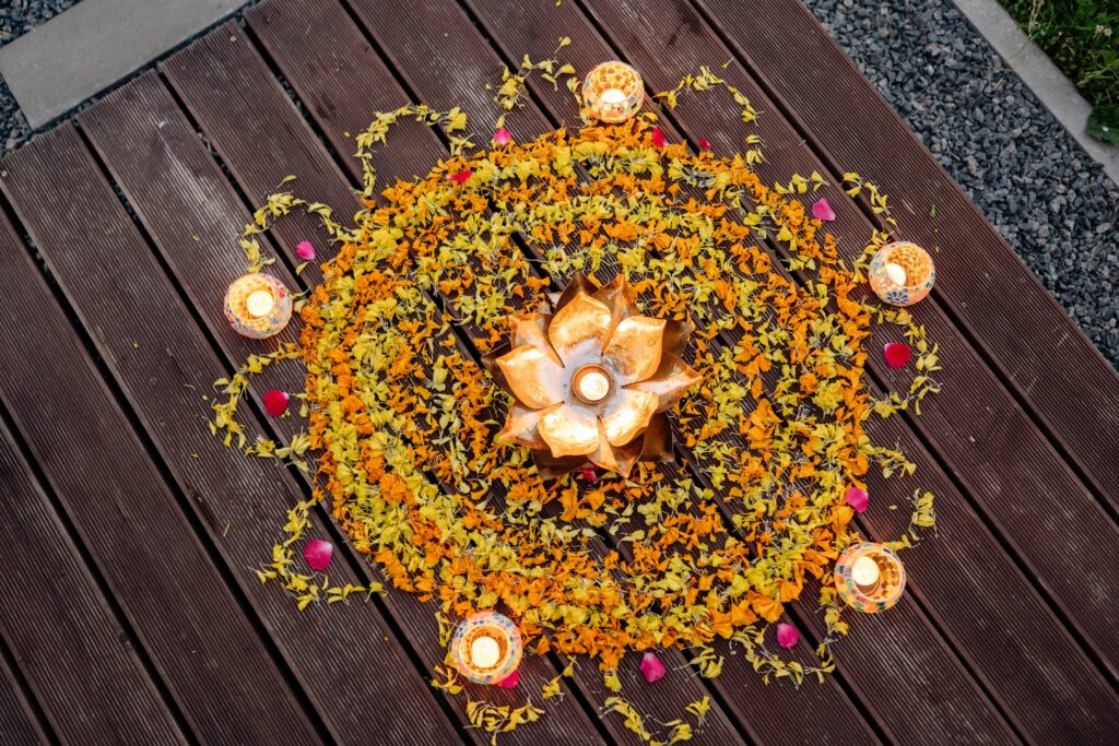 Vue du dessus d'une composition artistique sur un plancher en bois, formant un cercle de pétales de fleurs orange et jaunes (cempasúchil) entourant un bougeoir doré en forme de fleur, le tout illuminé par de douces bougies.