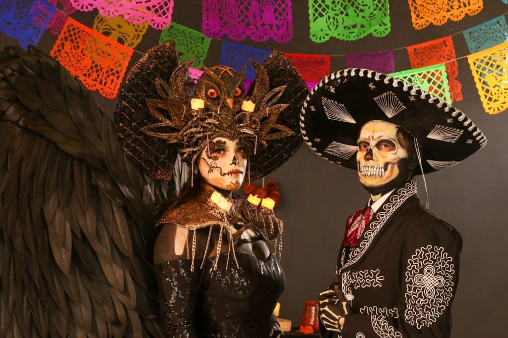 Portrait d'un couple posant en costumes et maquillages élaborés du Jour des Morts : une femme avec une coiffe florale ornée d'une chouette et un homme en tenue de charro, devant une arche de guirlandes de papel picado.