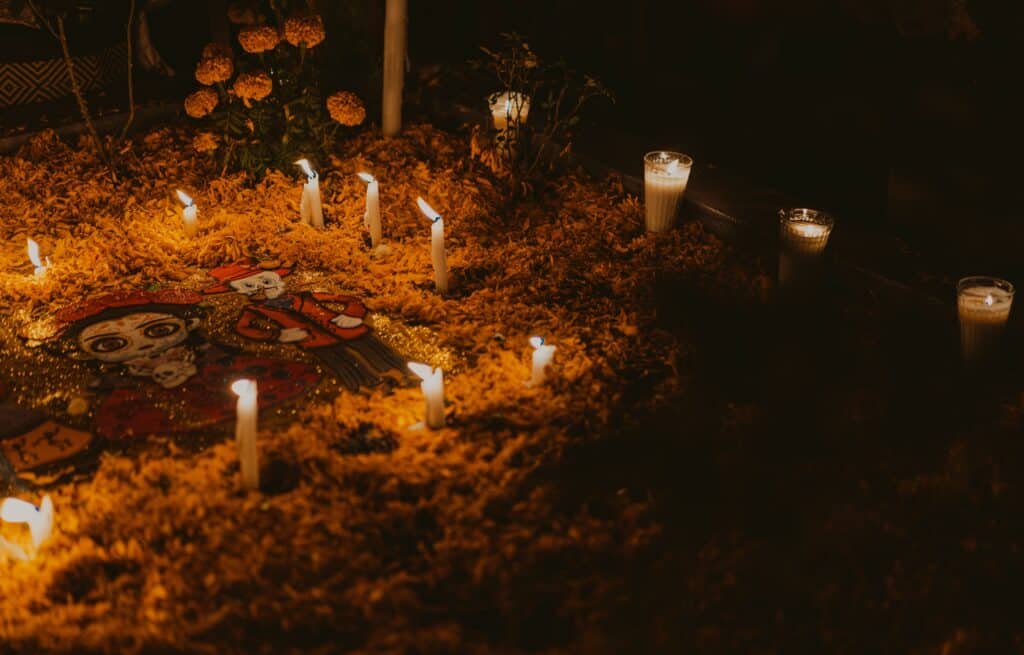 Ambiance nocturne dans un cimetière au Mexique pour la dìa de los muertos. Illuminé par de nombreuses bougies et recouvert de pétales de fleurs orange (cempasúchil). On distingue des objets d'offrande sur le sol.