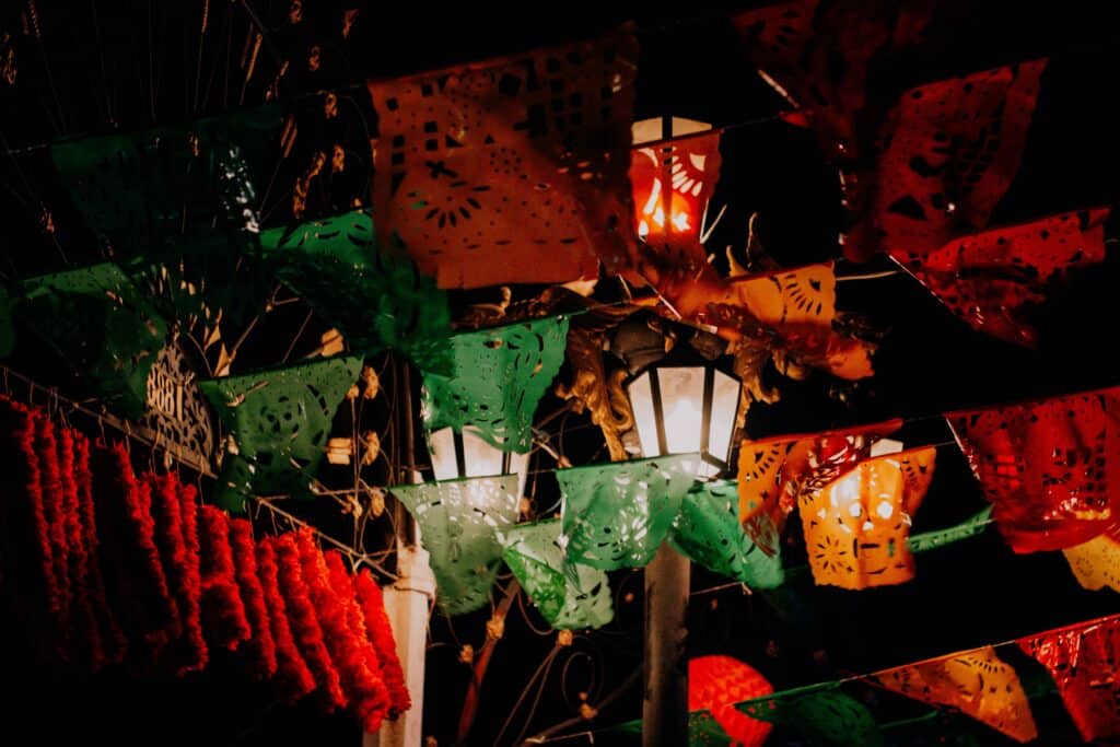 Gros plan de guirlandes de papel picado (papier découpé) suspendues dans l'obscurité de la nuit, illuminées par des lampions colorés créant une ambiance festive et tamisée.