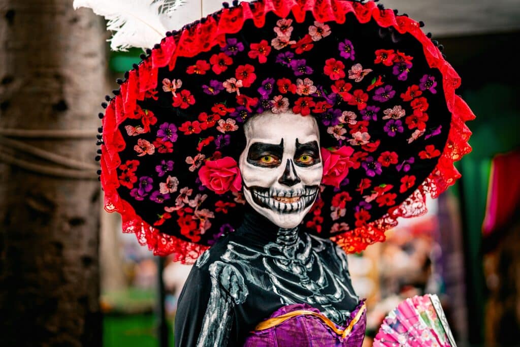 Gros plan d'une femme maquillée en Catrina élégante, portant un chapeau noir à larges bords orné de fleurs rouges et violettes, avec un costume squelette peint et un éventail rose à la main.