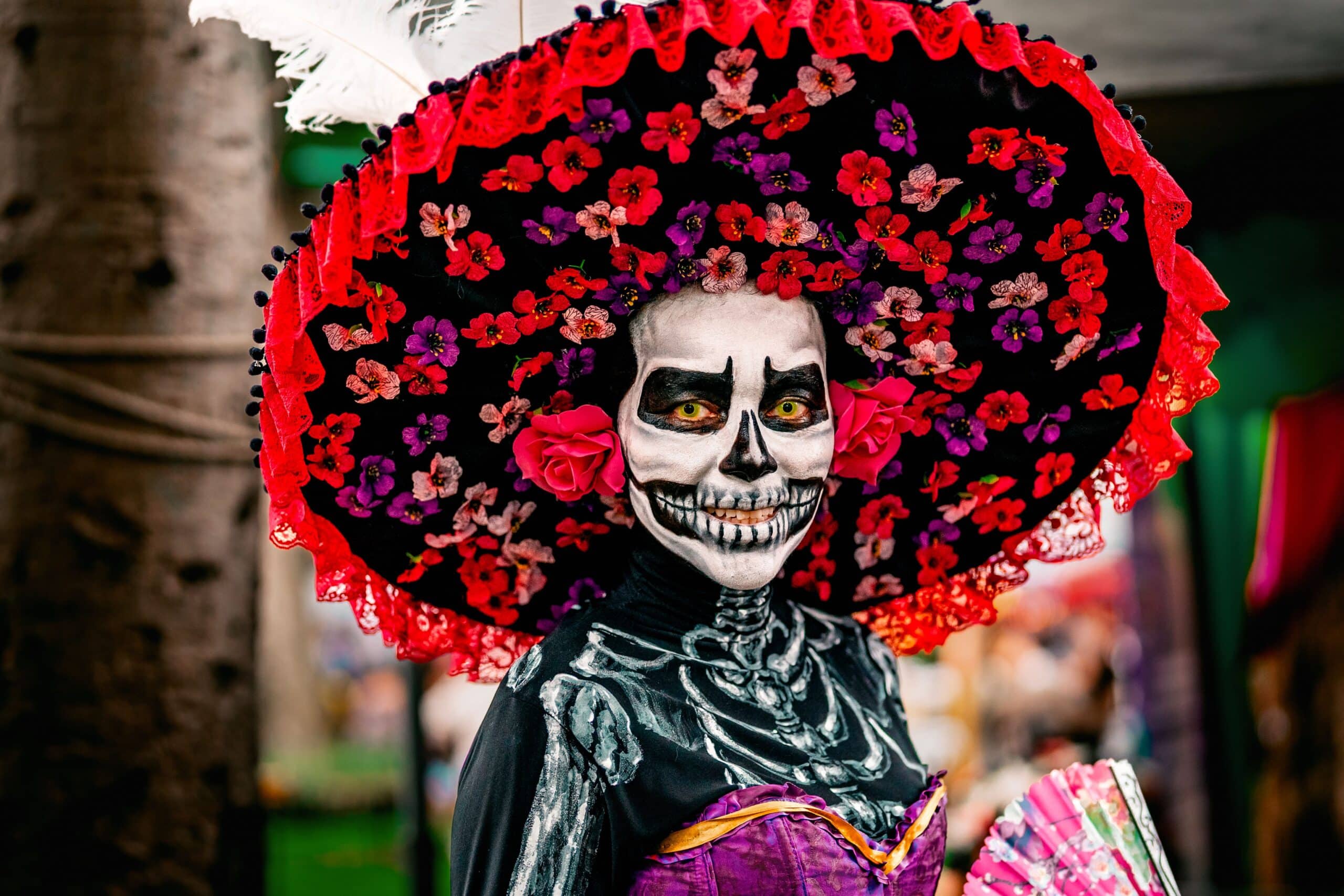 Gros plan d'une femme maquillée en Catrina élégante, portant un chapeau noir à larges bords orné de fleurs rouges et violettes, avec un costume squelette peint et un éventail rose à la main.