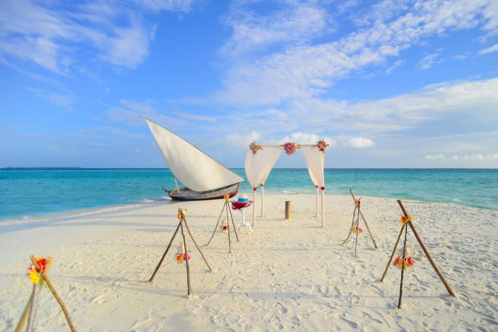 Décoration de mariage intimiste sur une plage de sable blanc au bord d'un lagon turquoise, avec une arche drapée de blanc, des petits trépieds en bois fleuris et un bateau à voile traditionnel à l'arrière-plan sous un ciel bleu lumineux.