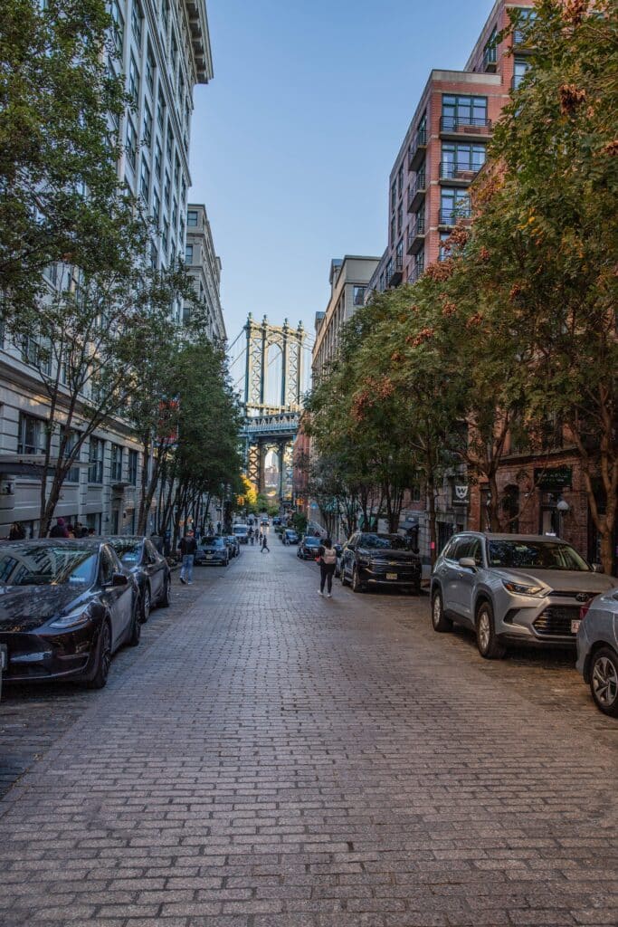 Rue pavée pittoresque bordée d'arbres et de briques rouges avec vue sur le pont de Brooklyn, l'âme créative de New York par Heaven Collection.